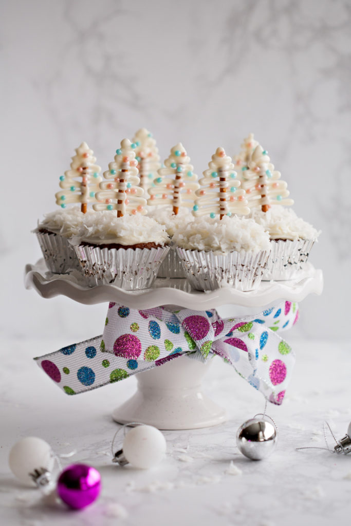 Festive Gingerbread Cupcakes with Cream Cheese Frosting and cute pretzel Christmas tree toppers on a bed of fluffy coconut!